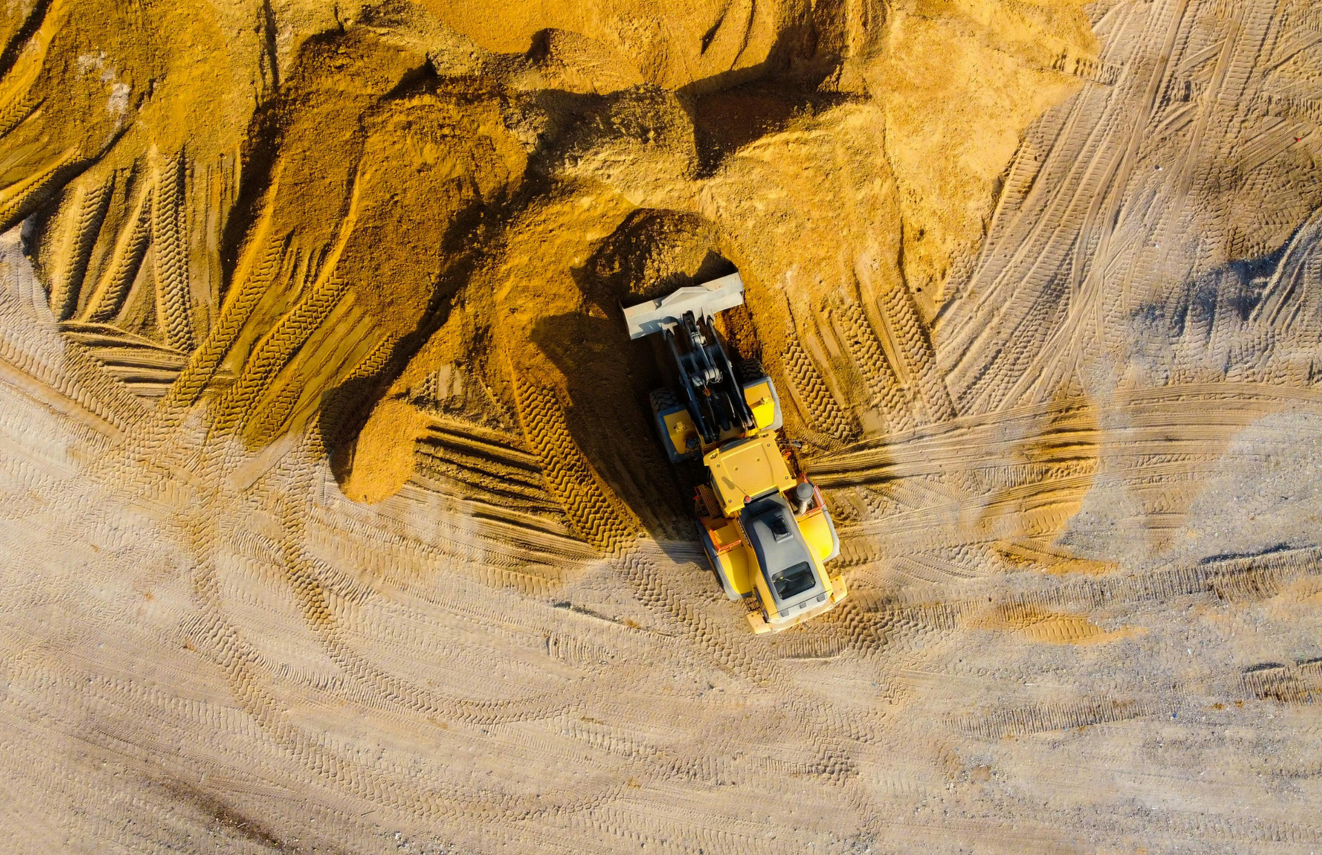 Yellow loader working on a pile of golden-brown material, leaving tracks in a sandy area.