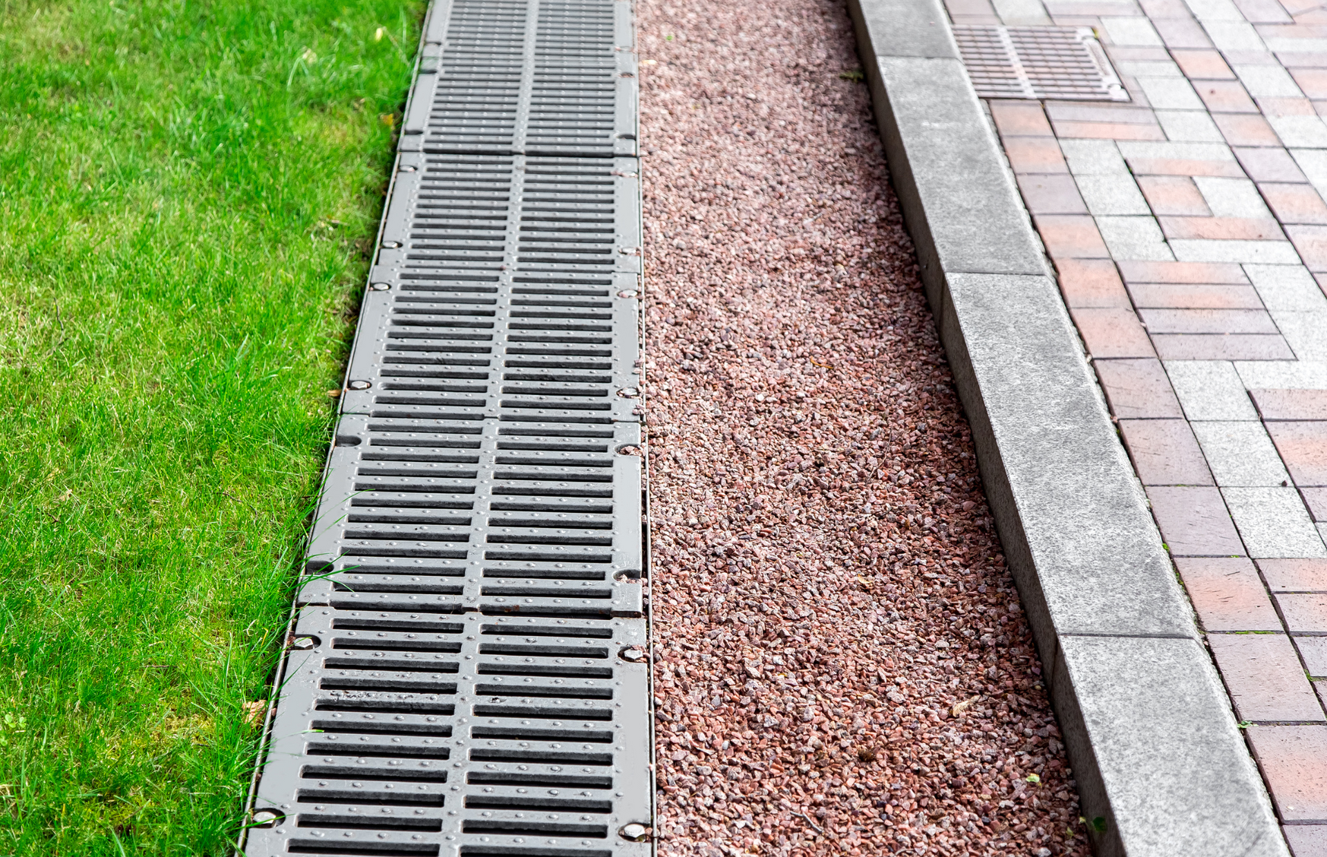 Drainage grate between grass and brick sidewalk, with gravel bed.