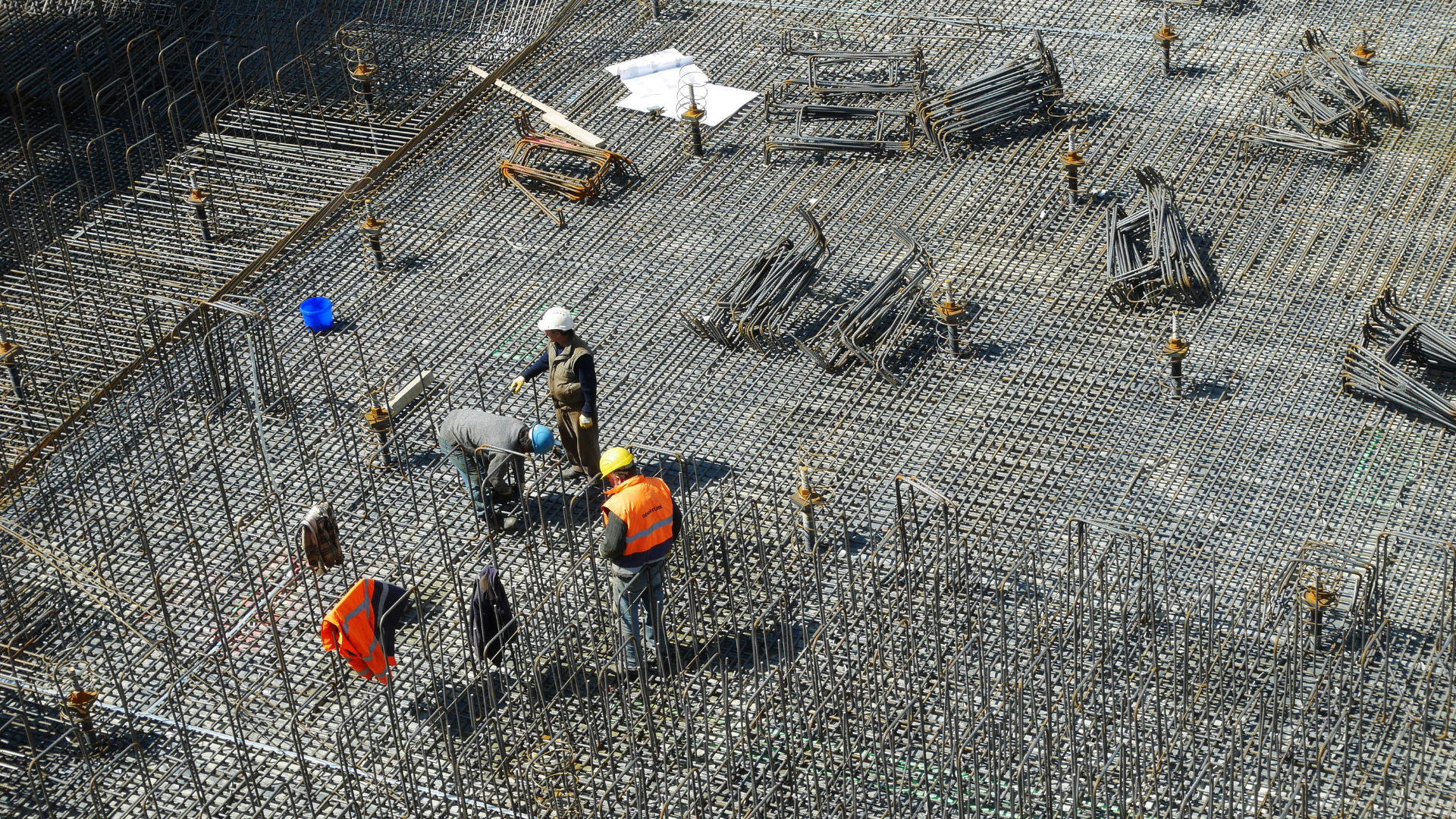 Construction workers on a site with rebar. One in orange vest and hardhat points while others work.