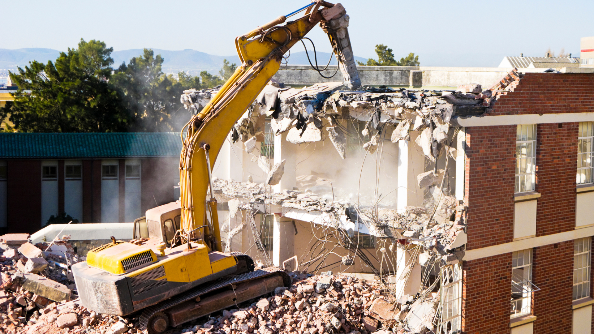 Yellow excavator demolishing a multi-story brick building on a sunny day.