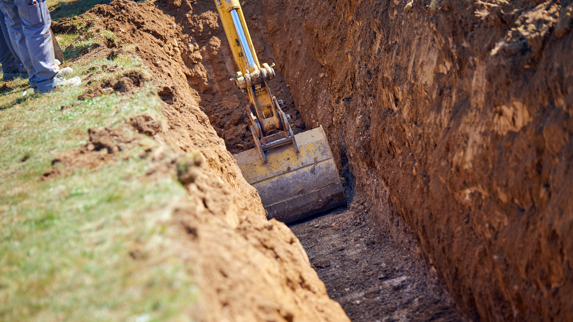 Excavator digging a trench in brown soil, with a person standing nearby on green grass.