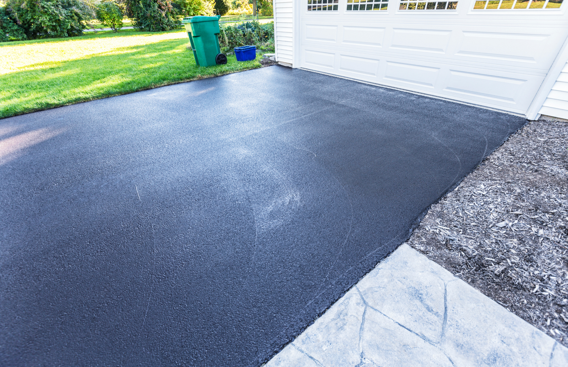 Freshly sealed black asphalt driveway with green grass, white garage, and concrete path.