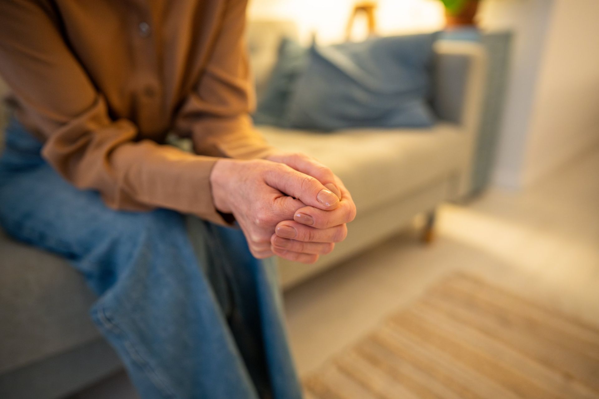 Person sitting on a couch, hands clasped, brown shirt, blue jeans, neutral setting.