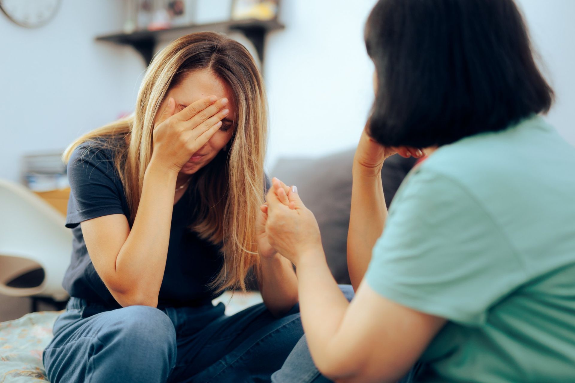 Woman crying with hand over face, comforted by another woman's hand. Sitting on a couch.