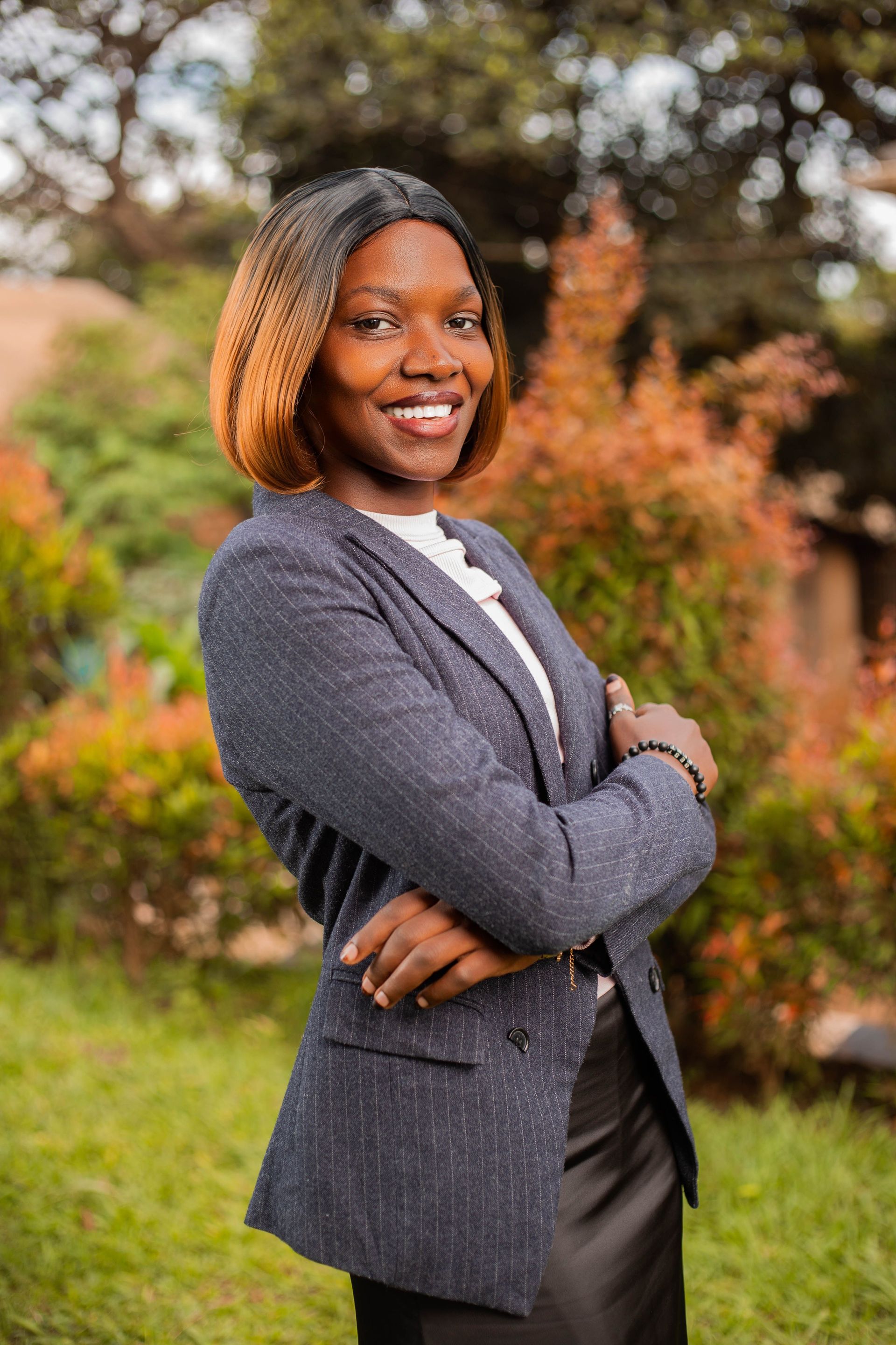 Smiling person in a gray blazer standing outdoors with arms crossed, in front of autumn foliage.