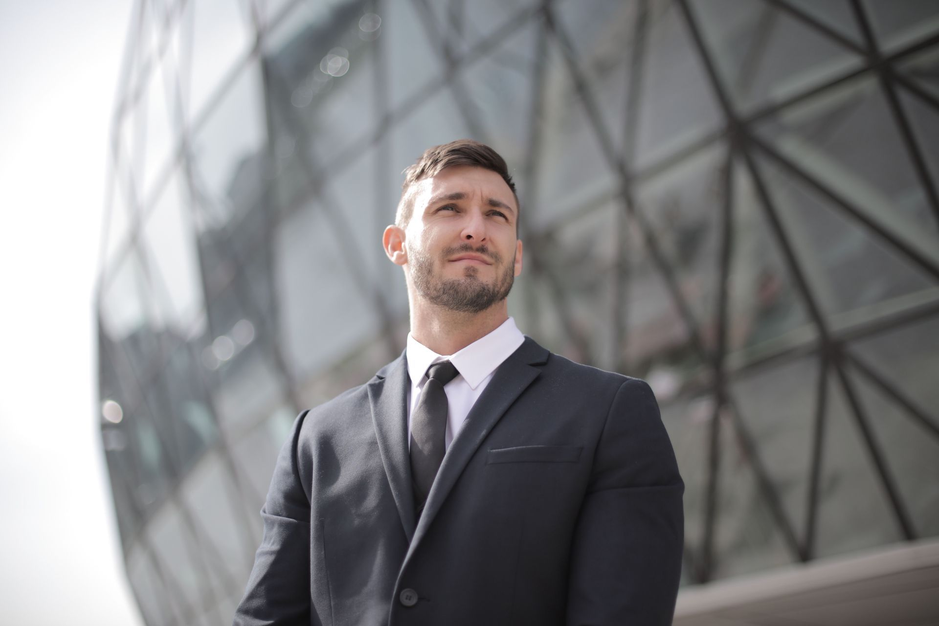 Man in a dark suit standing outdoors before a glass building, looking off to the side.