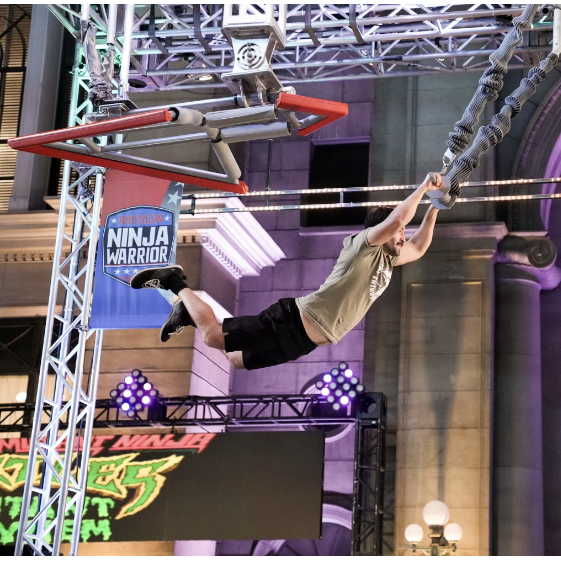 A man is hanging upside down in front of a sign that says ninja warrior