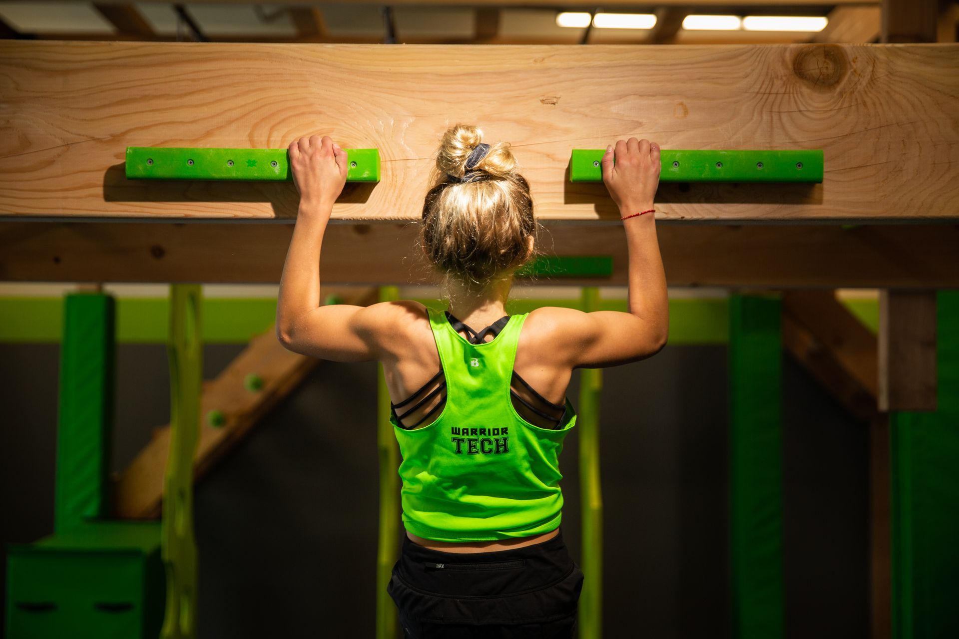 A woman in a green tank top is doing pull ups on a wooden bar.
