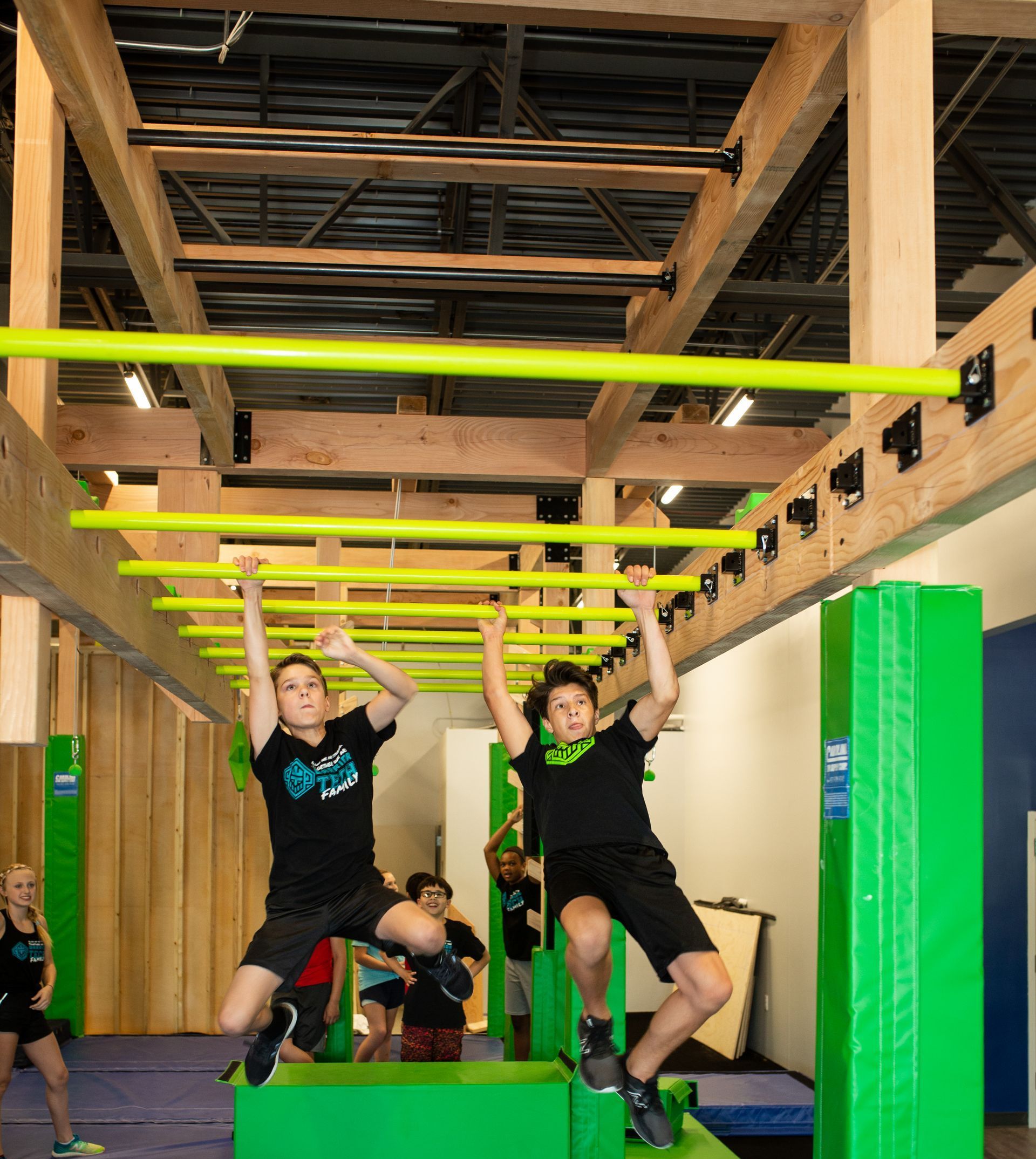 Two people hang from a yellow overhead obstacle course, green blocks below.