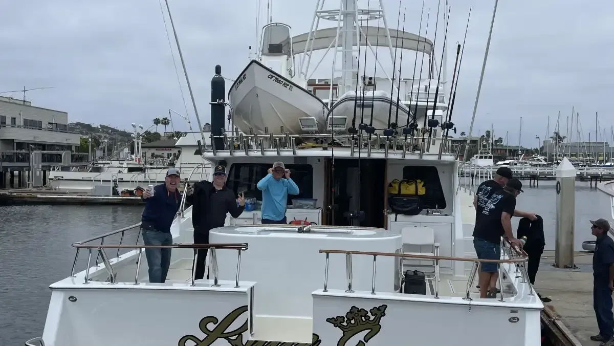 People on a white boat docked at a marina, with one person waving and a large ship in the background.