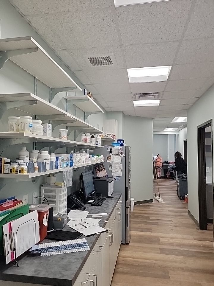 Medical supply room with shelves of bottles, a workstation with a computer, and a hallway leading to an office area.