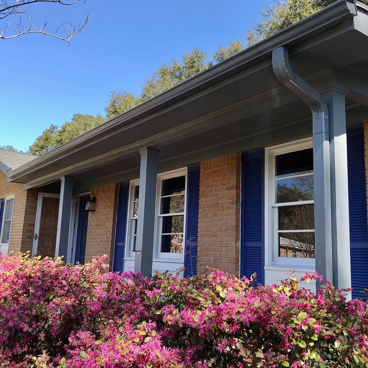 A brick house exterior with a porch, blue shutters, grey trim, and blooming pink azalea bushes in the foreground.