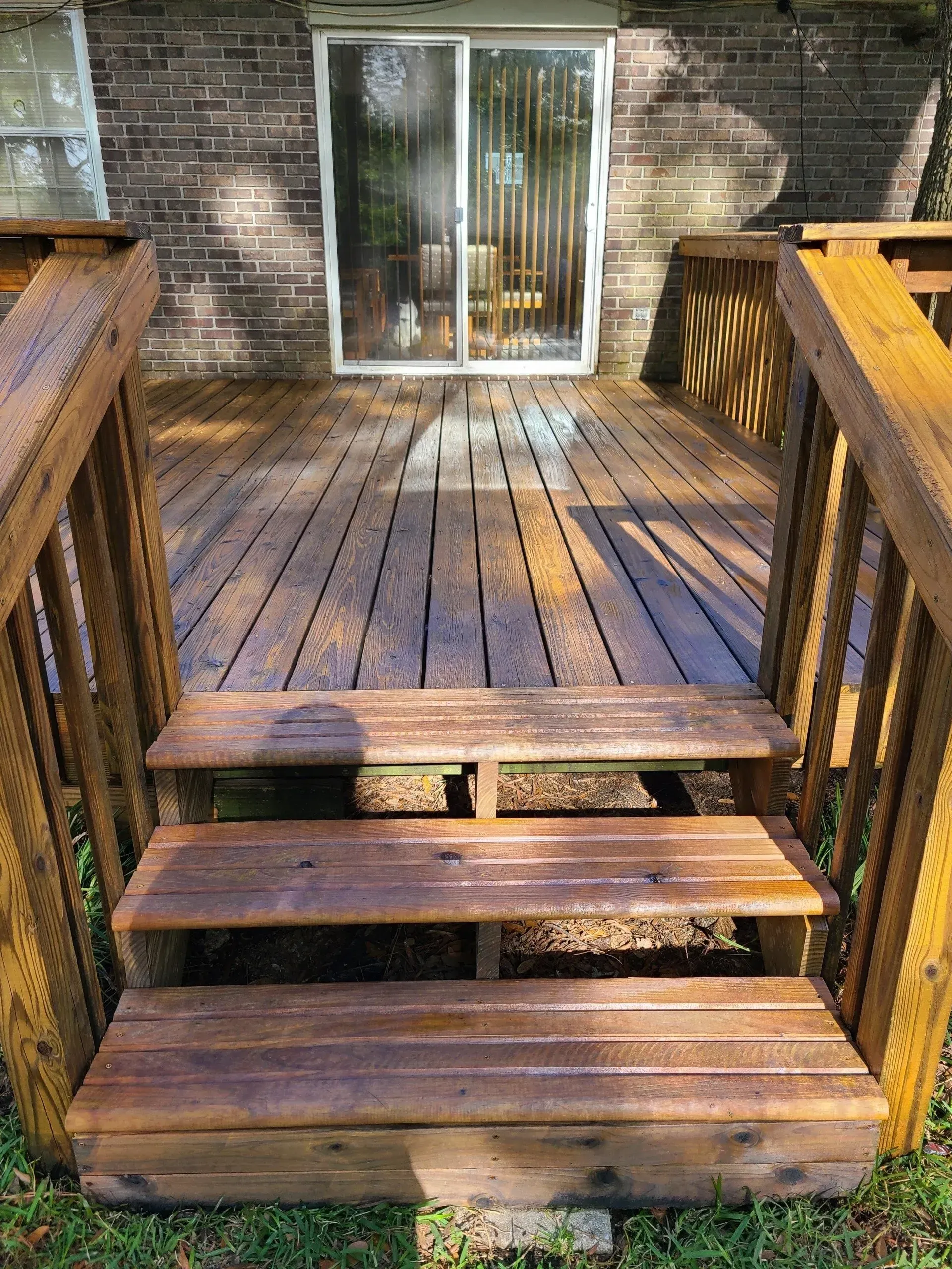 A wooden deck with a set of three stairs leading up to a sliding glass door on a brick house.