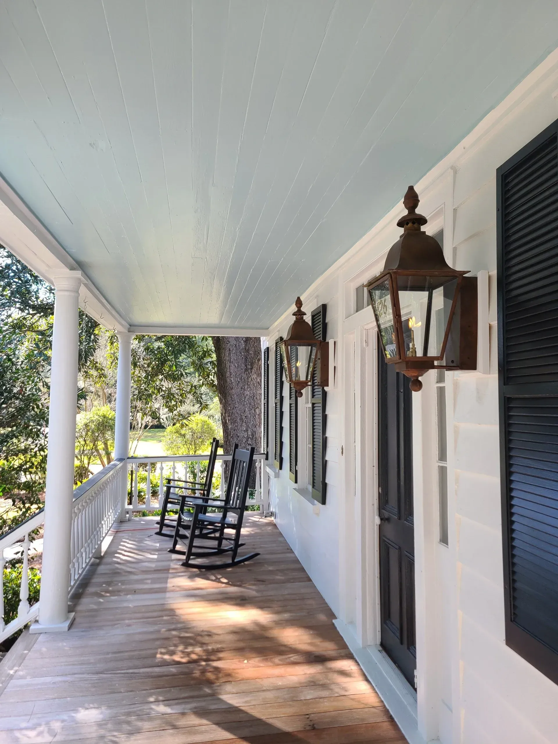 A white porch with blue ceiling, black shutters, antique-style wall lanterns, and two wooden rocking chairs.