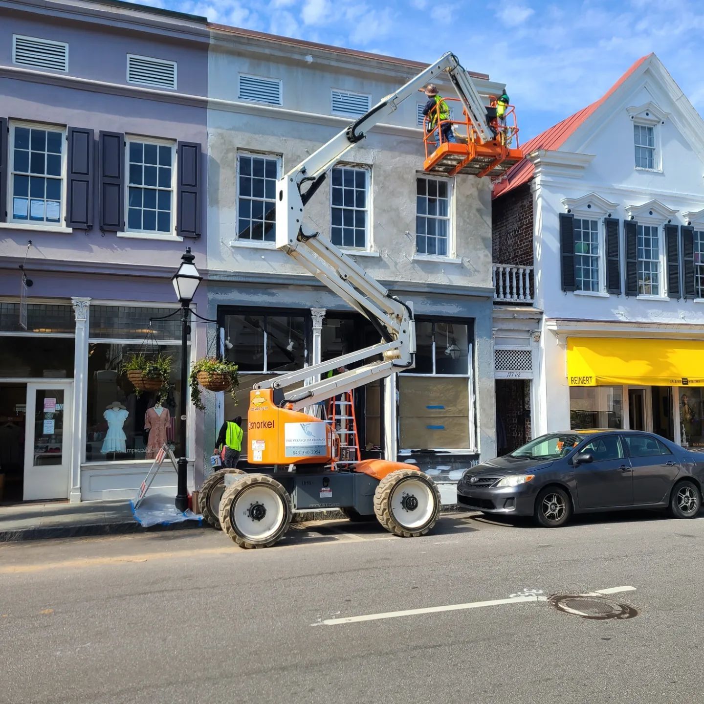 A cherry picker lift reaches the facade of a light-colored commercial building where two workers perform maintenance.