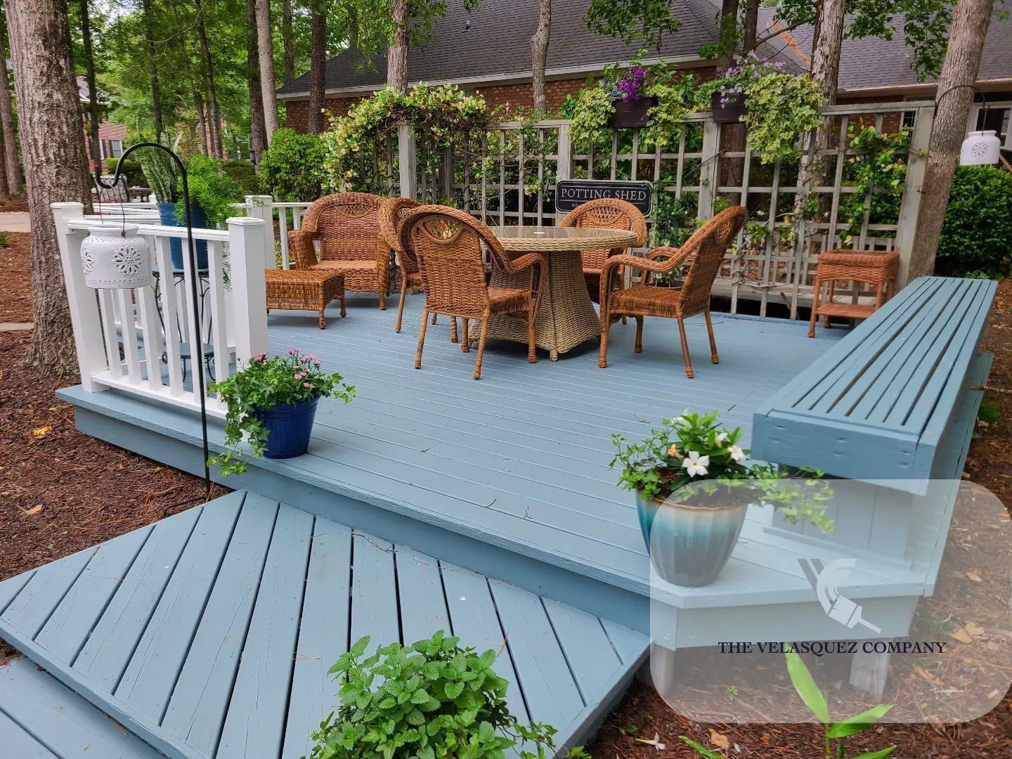 Blue painted deck featuring a dining table with wicker chairs, potted plants, a wooden bench, and white railings.