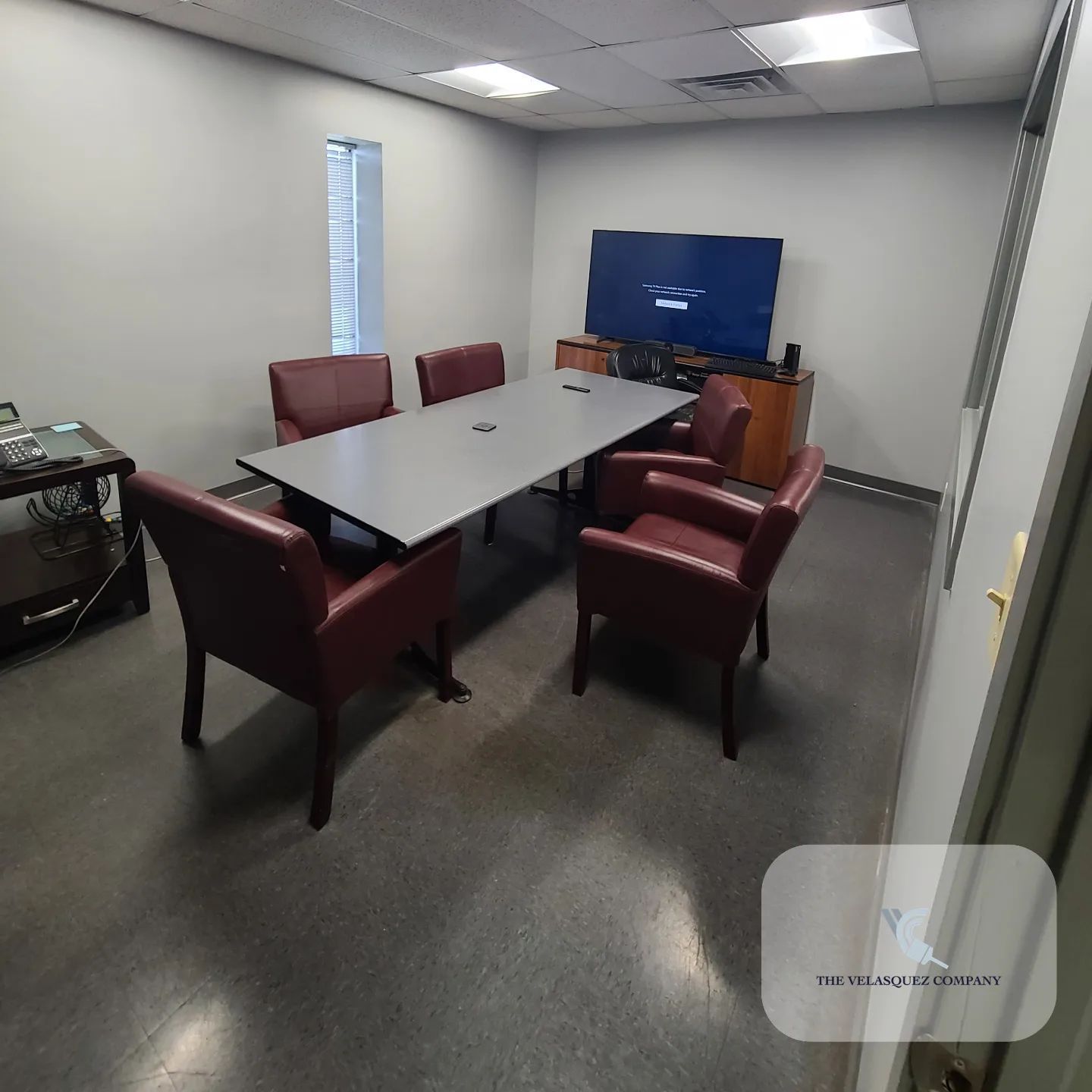 A conference room with a gray table, six dark red chairs, and a wall-mounted TV on a wooden stand.