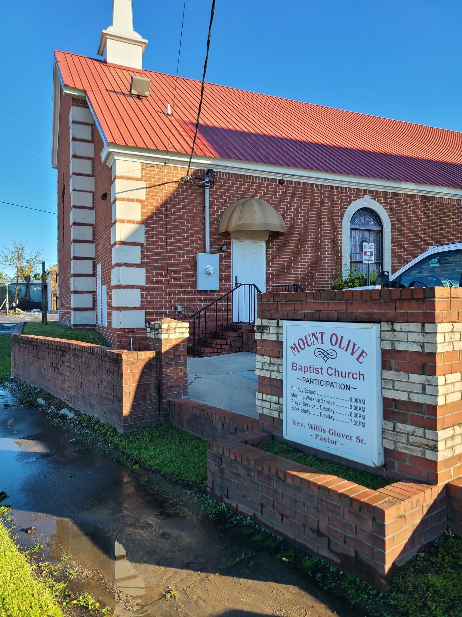 A red brick church building with a white steeple and a sign out front on a sunny day.