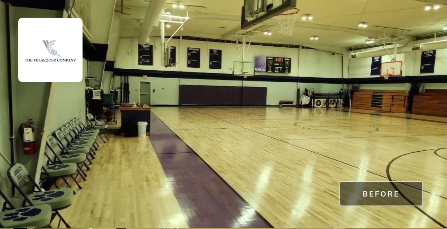 An empty basketball court with wooden floors, purple borders, bleachers, and a row of chairs along the sideline.