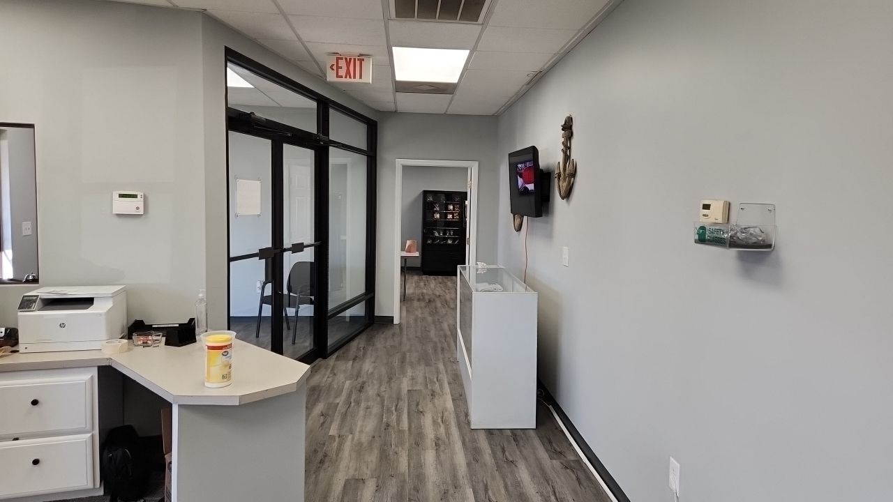 An office hallway with grey walls, light wood-grain floors, a reception desk, a glass-walled office, and a wall-mounted TV.