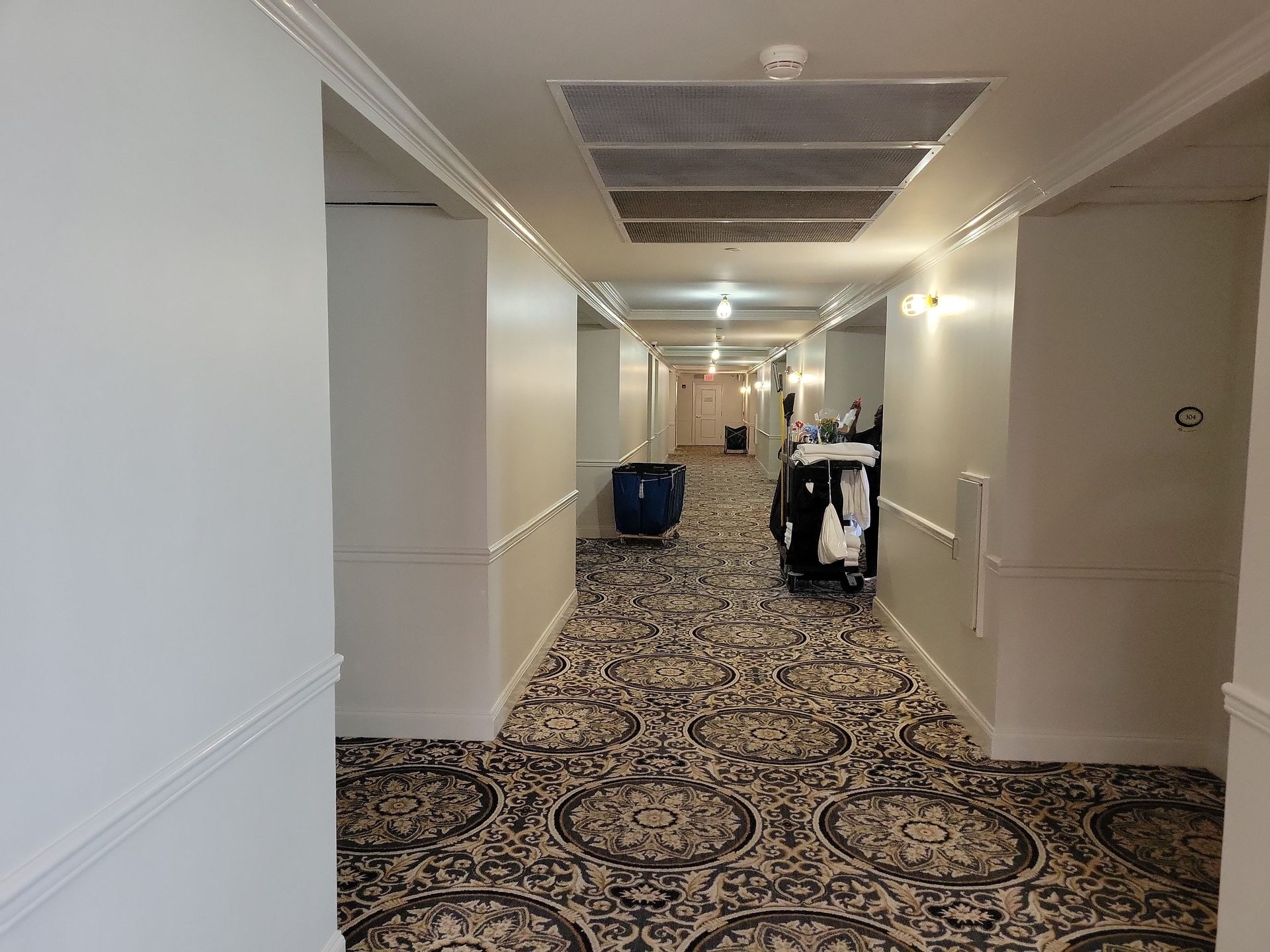 A long hotel hallway with white walls and a patterned carpet, featuring a housekeeping cart parked on the right.
