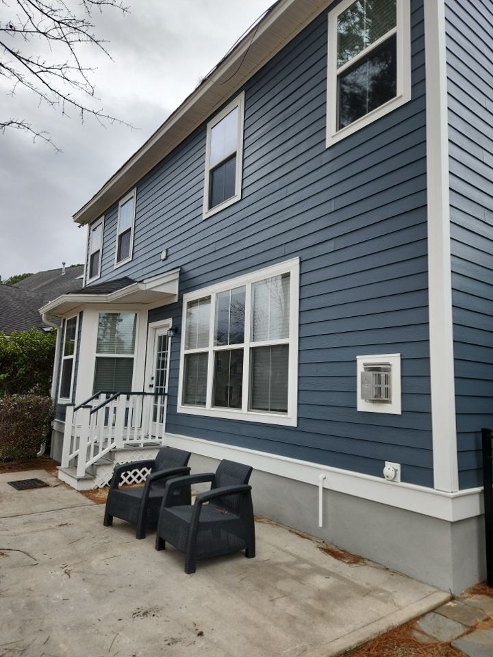 Two black patio chairs sit on a concrete patio next to a two-story blue house with white trim.