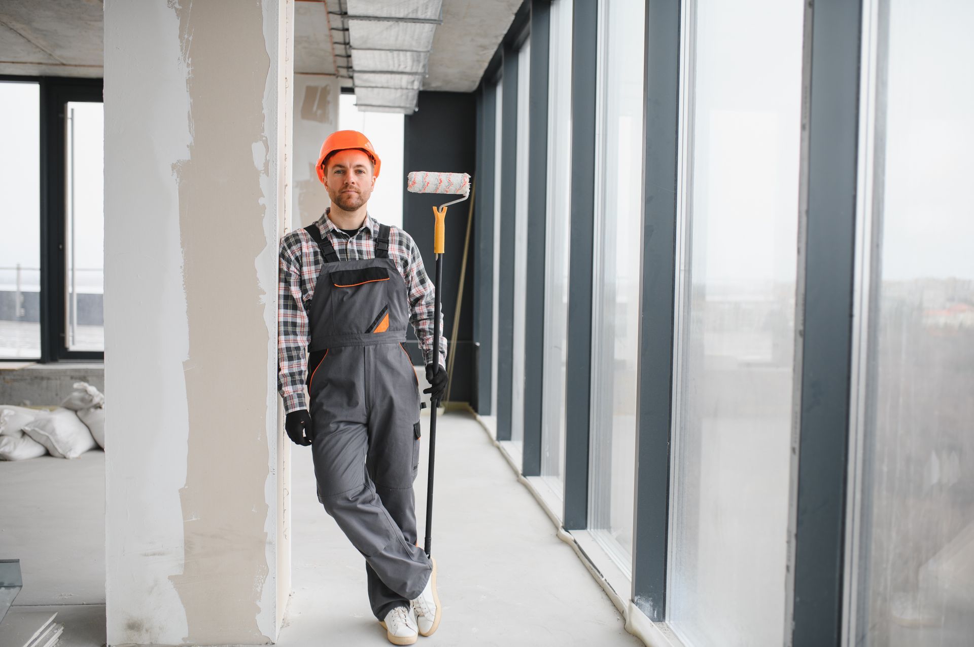 A male worker holding a paint roller near a window in a commercial building under construction. A painter wearing a hard hat and grey overalls leans against a pillar in an unfinished room with large glass windows.