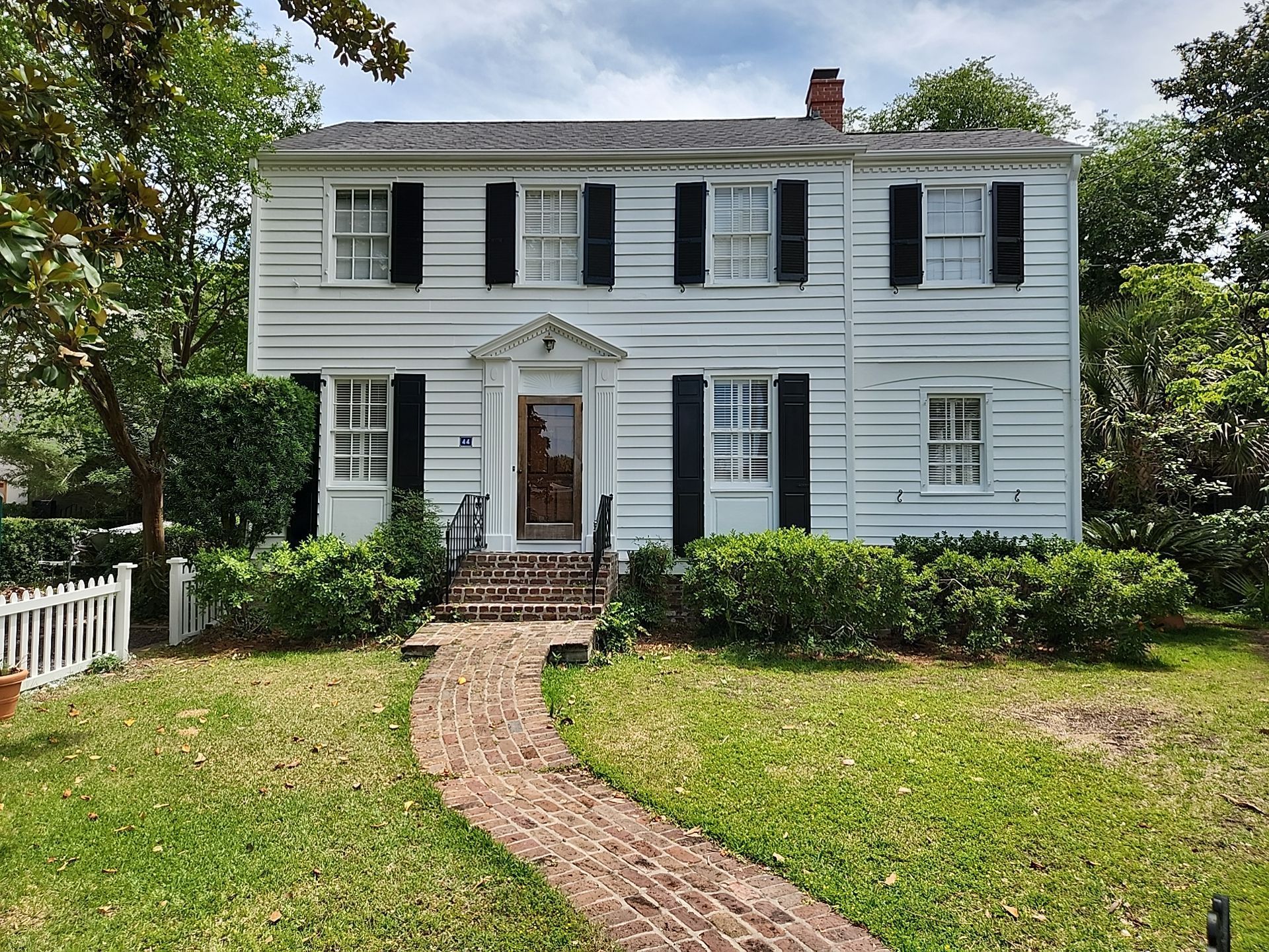 A two-story, white clapboard historic house with black shutters and a brick walkway leading to the front door.