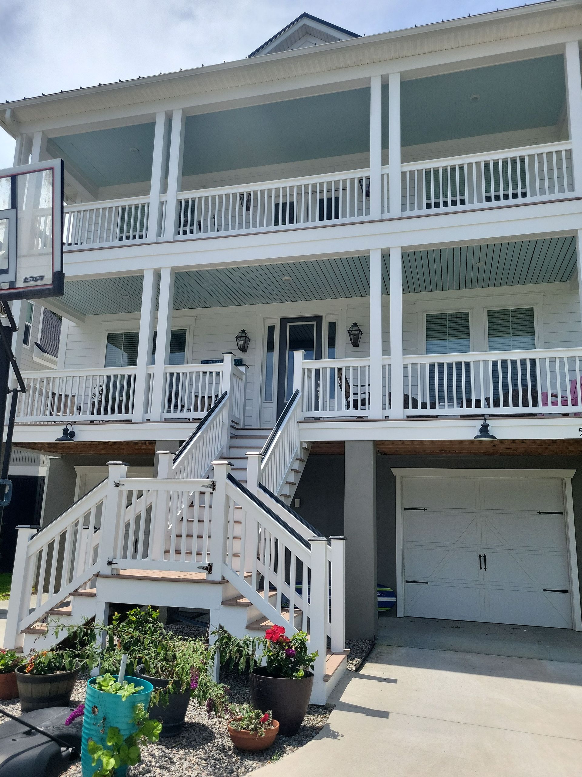A three-story, elevated white house with large front porches, a white staircase, and potted plants in the foreground.