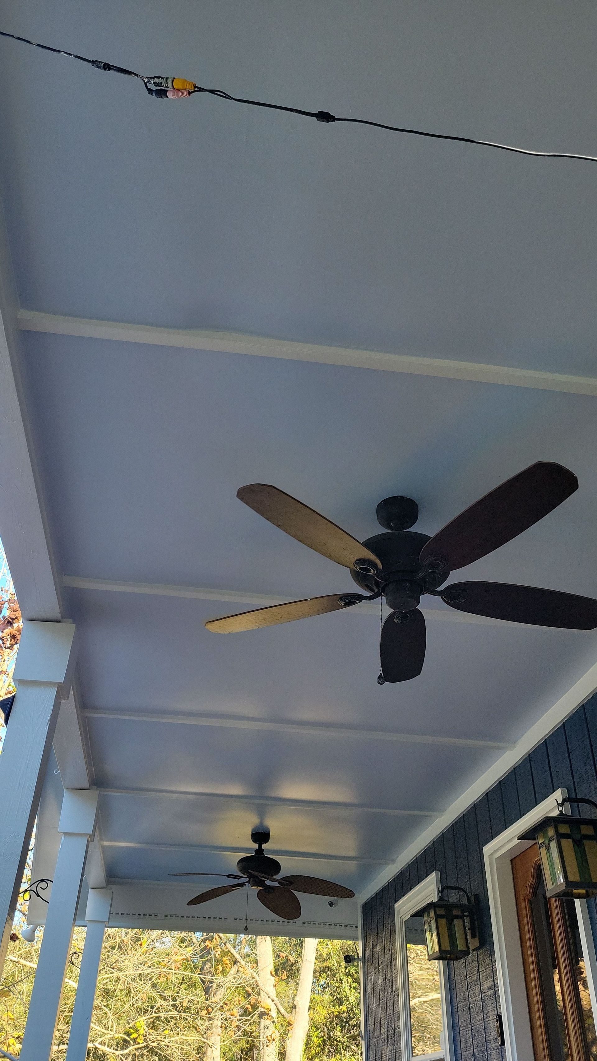 Two dark-colored ceiling fans hanging from the light-blue, slatted ceiling of a covered porch.
