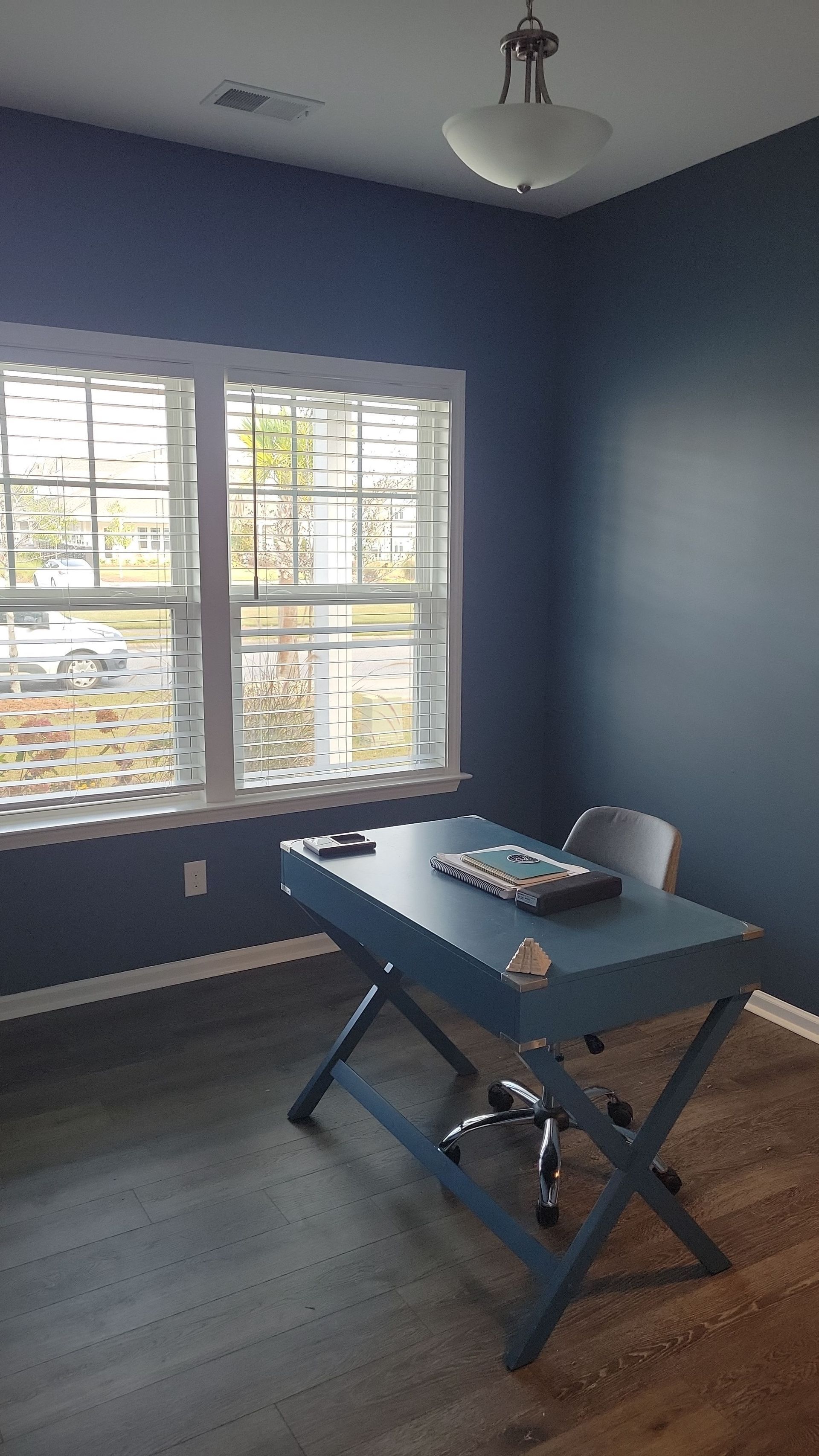 A blue desk and chair sit in a room with dark blue walls, wood flooring, and a large window with white blinds.