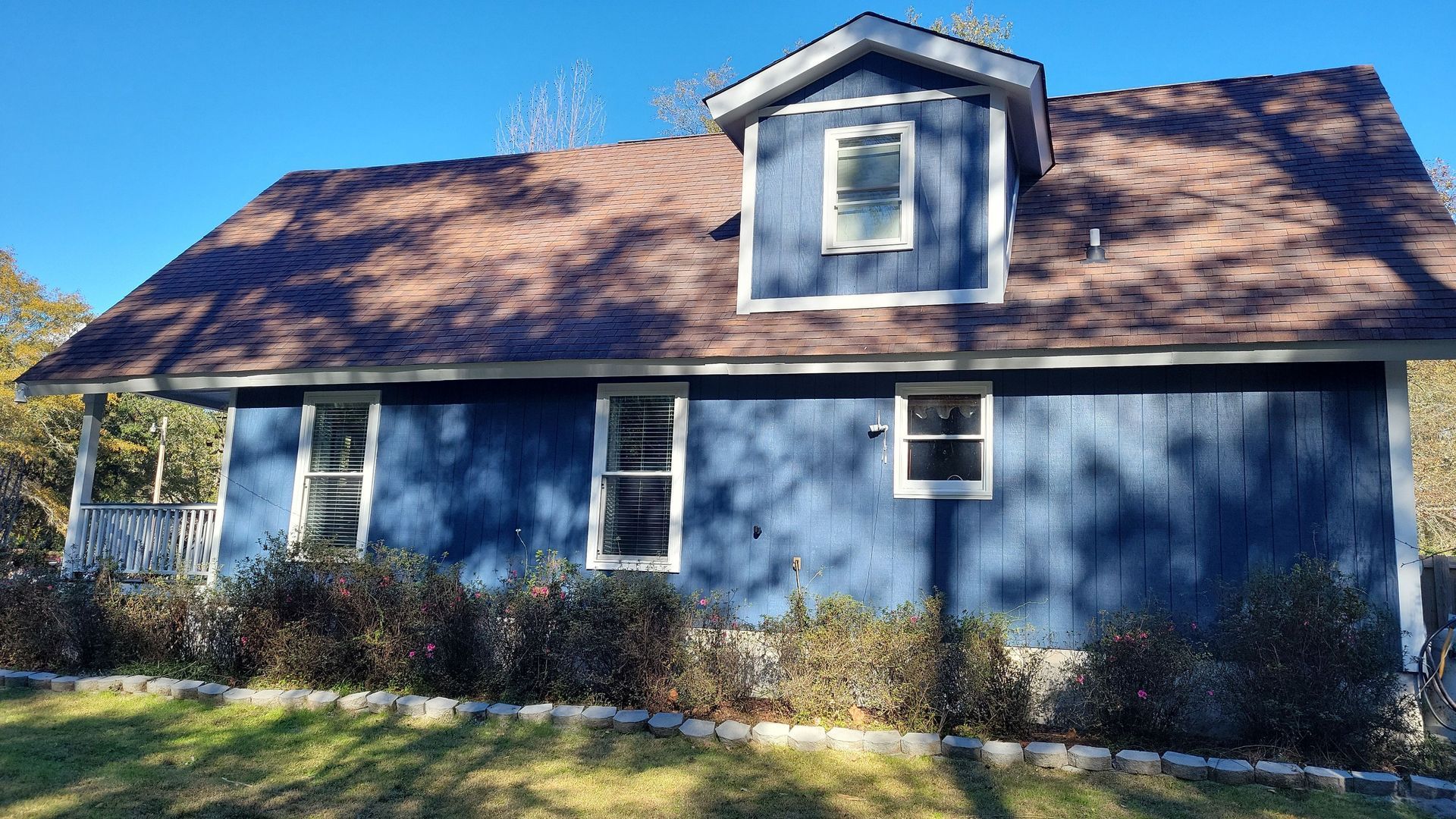 A blue, two-story house with a dormer window, brown shingles, and a white porch, surrounded by shrubs on a sunny day.