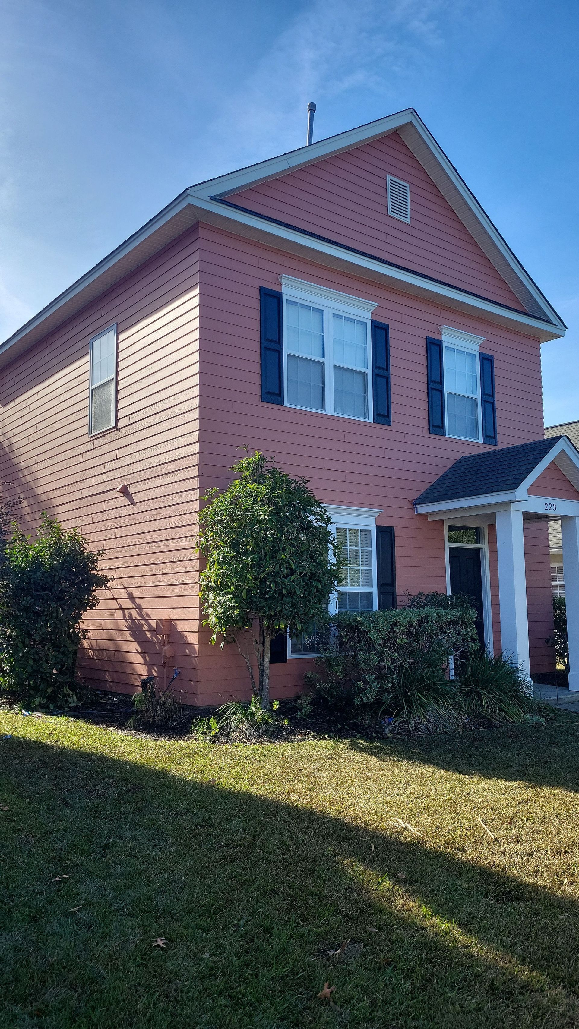 A two-story, pink, brick-sided house with a grey roof, dark shutters, and small bushes in the front yard.