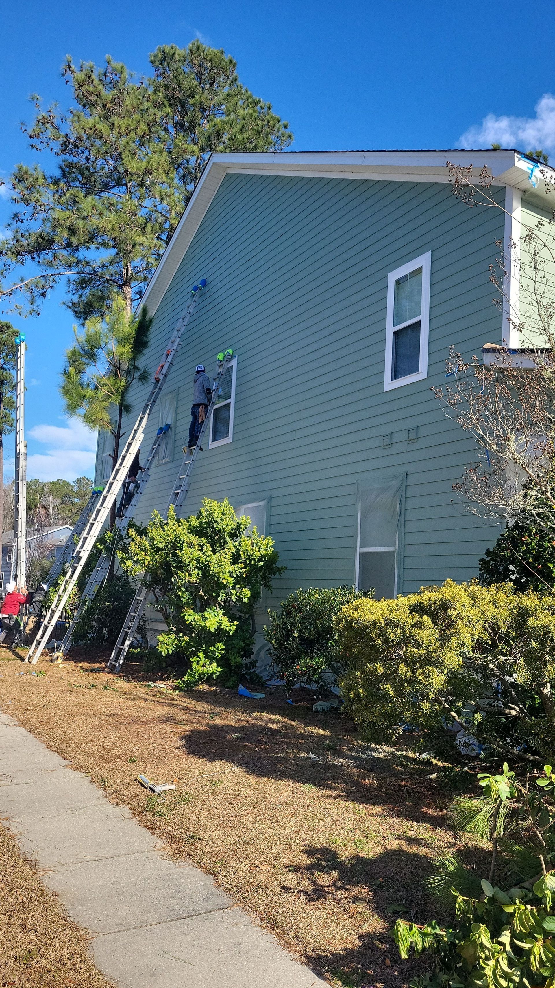 Two people on tall ladders work on the light green siding of the side of a house.