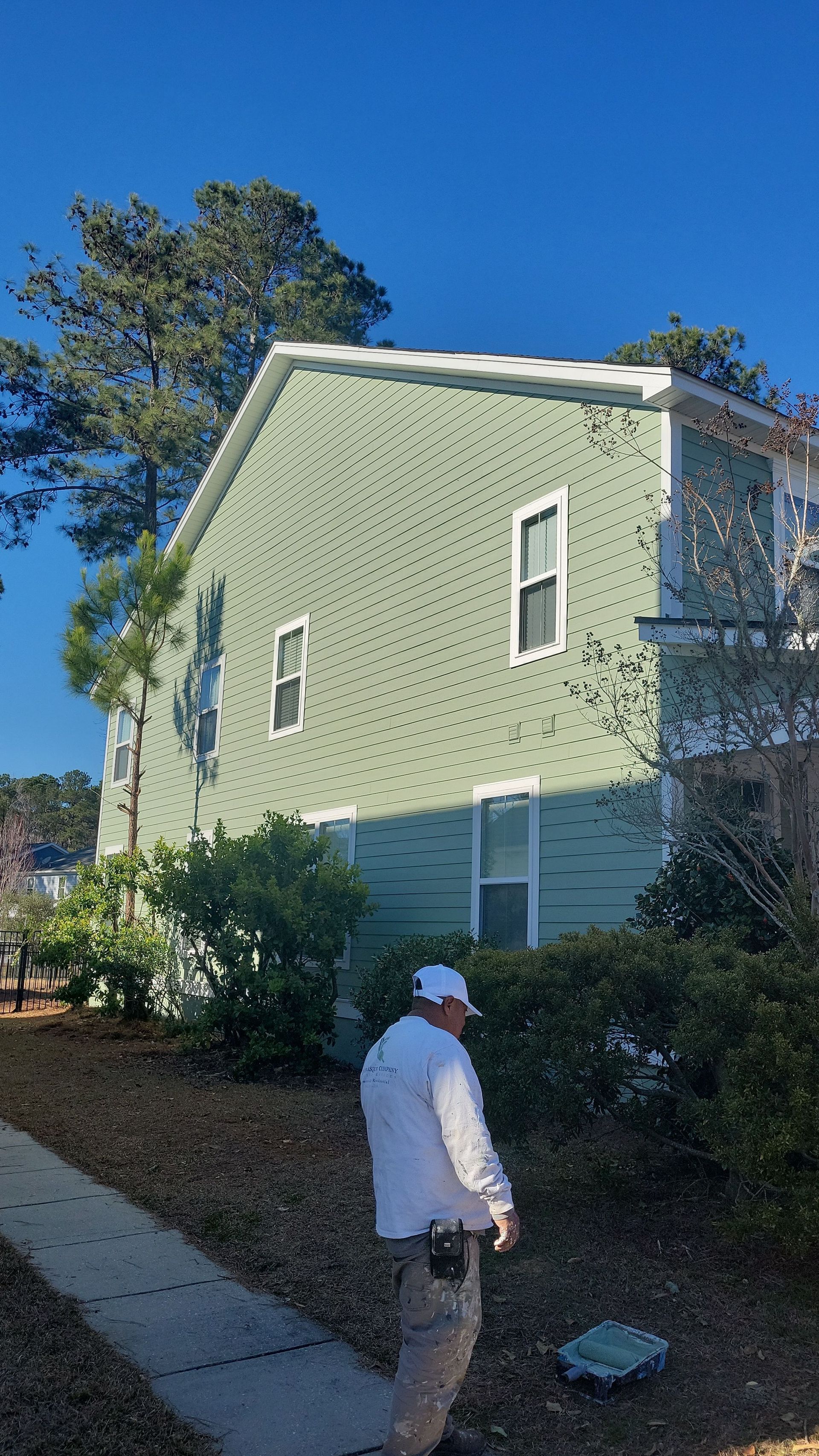 A person in a white shirt and light pants stands near a green-sided house and a sidewalk on a sunny day.