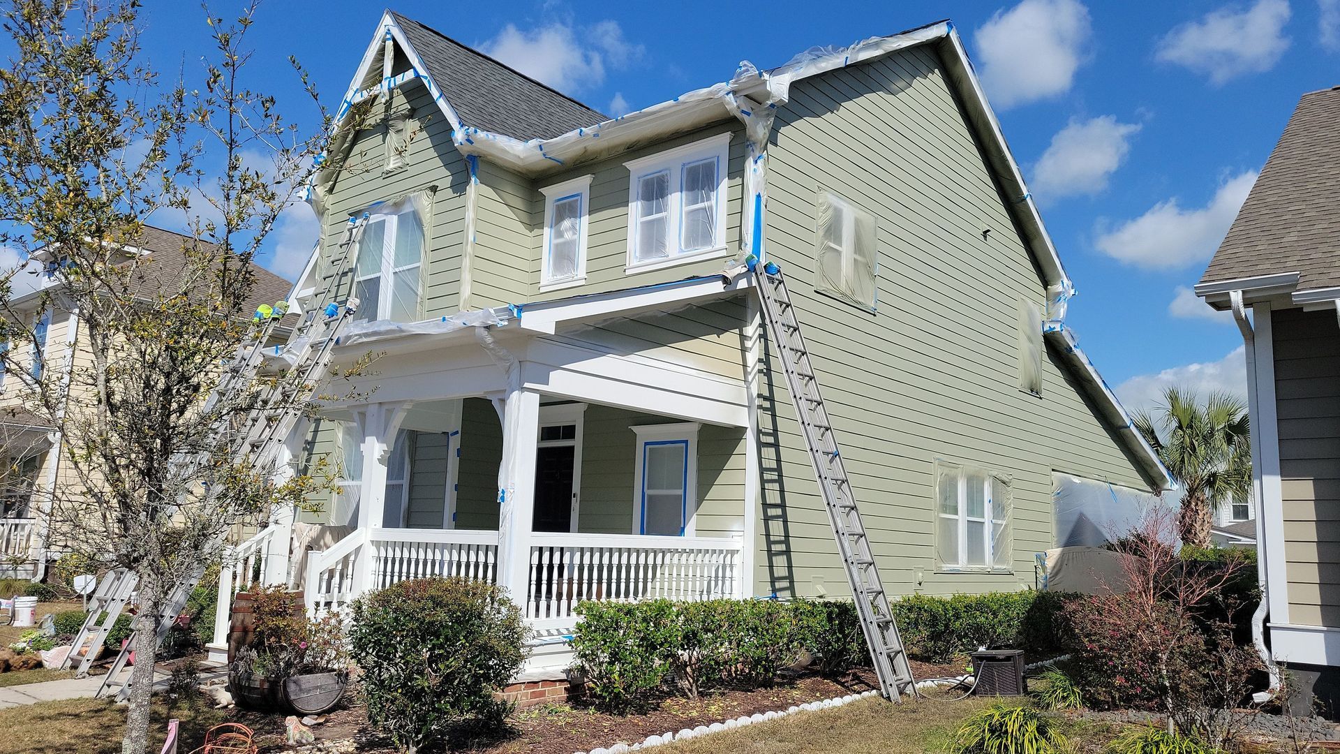 A light green two-story house with a white front porch and a ladder leaning against the side for exterior repairs.