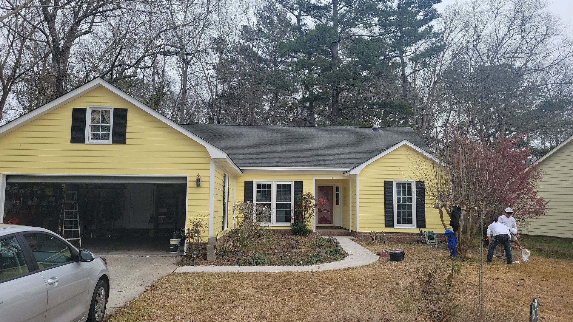 A yellow, single-story house with a garage and black shutters, while workers clear the yard during an overcast day.