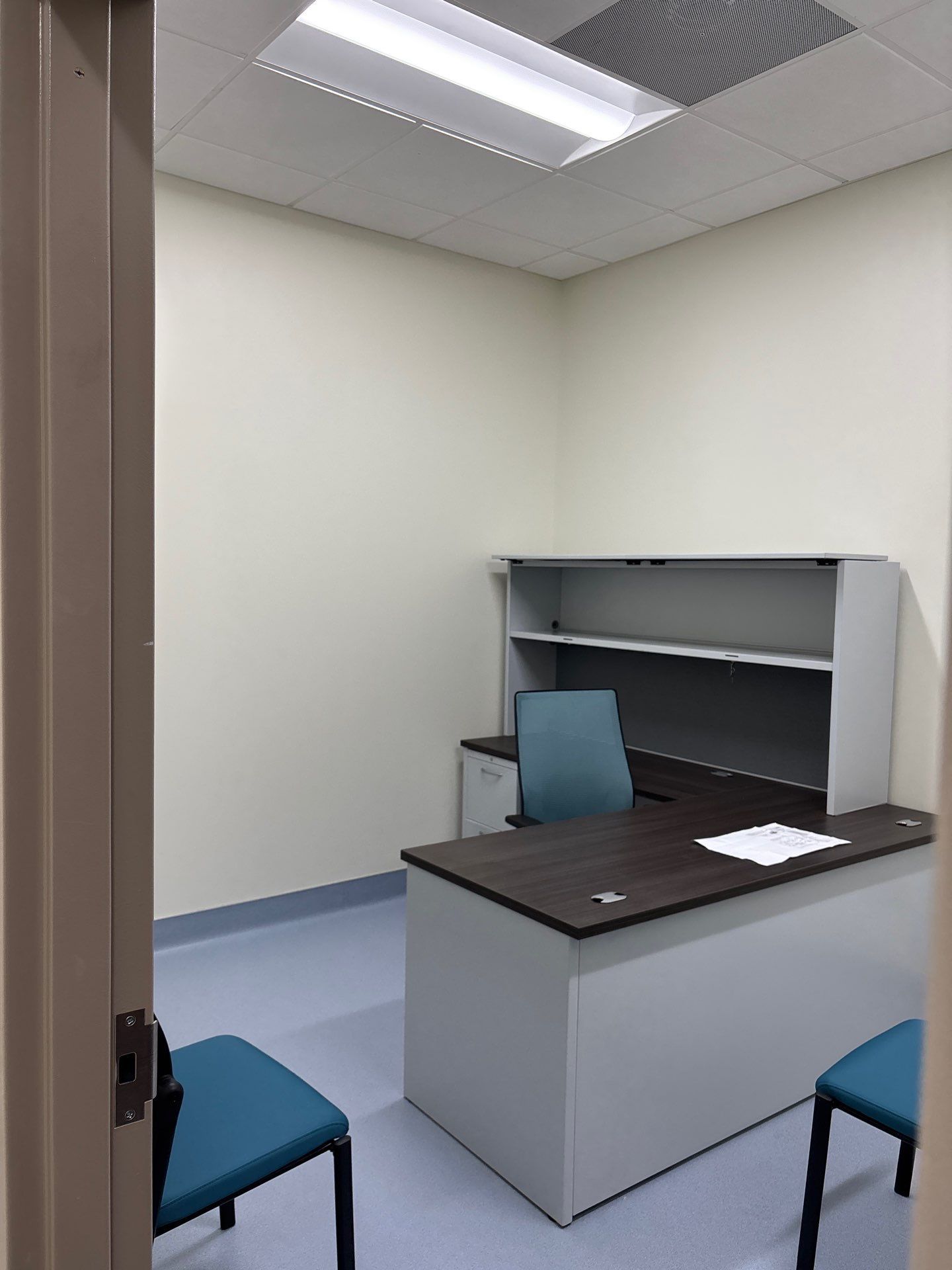 A small office with a dark wood desk, a teal office chair, two guest chairs, and a grey hutch against a white wall.