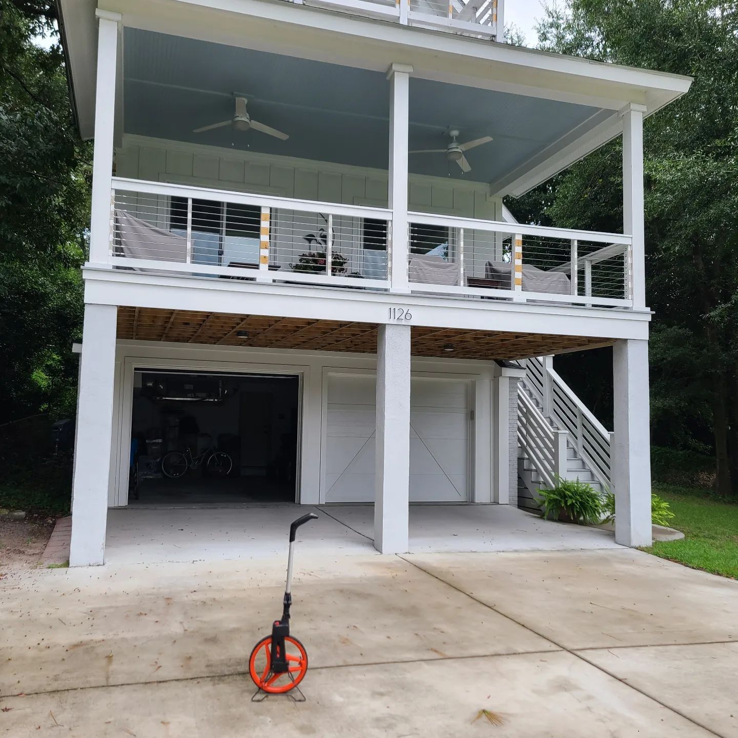 An elevated two-story house with a white exterior, garage space, and a balcony, with a measuring wheel on the driveway.