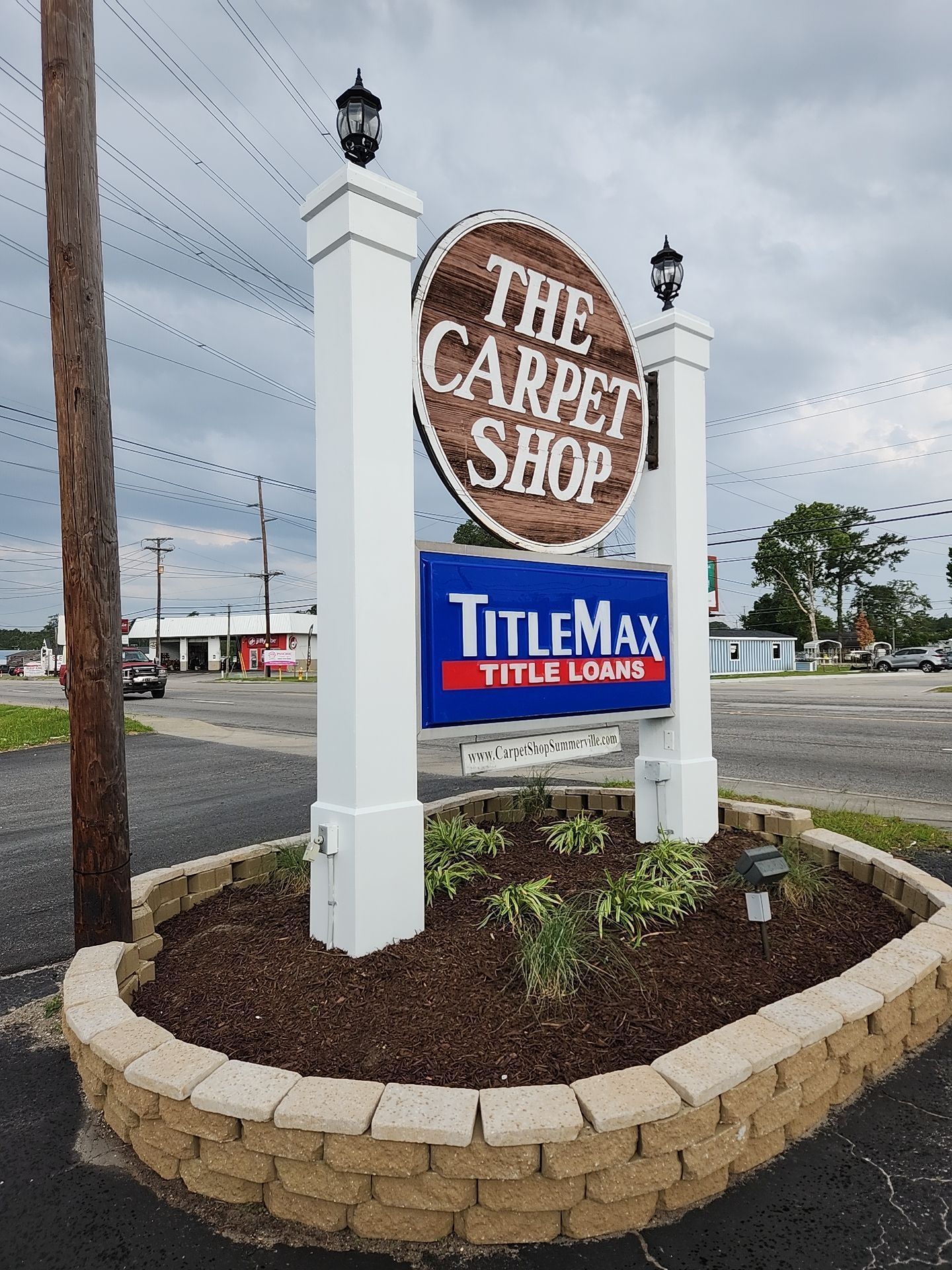 A street sign for The Carpet Shop and TitleMax Title Loans, set in a stone garden bed on a cloudy day.