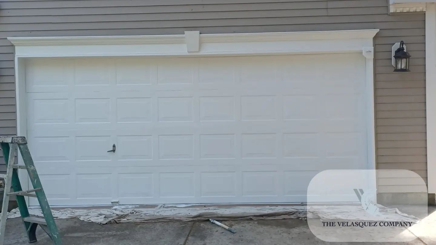A white garage door with decorative trim framing, set in a light brown siding wall with a ladder nearby.