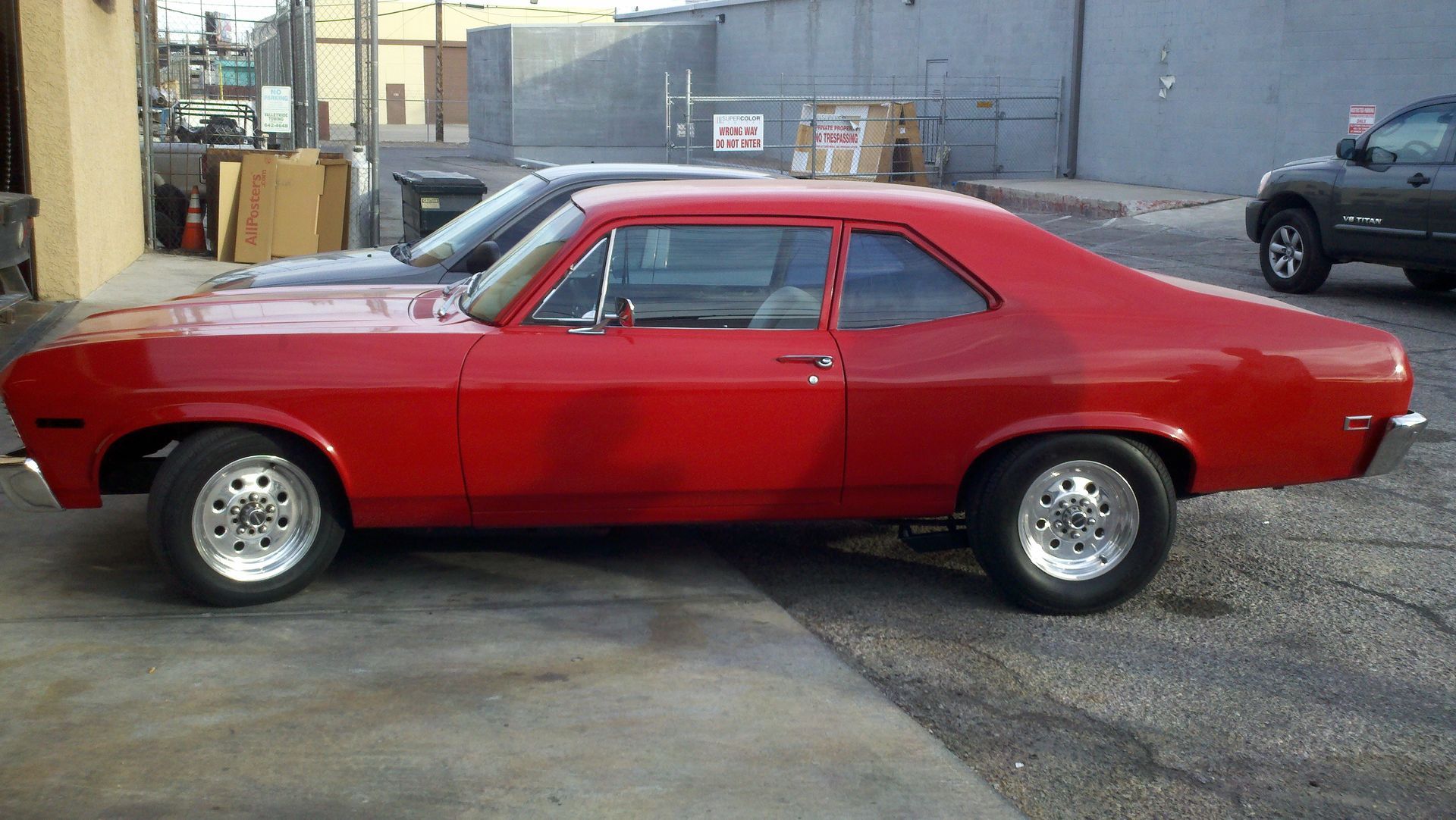 Red classic coupe parked on an industrial lot, side view, with chrome wheels and a glossy finish