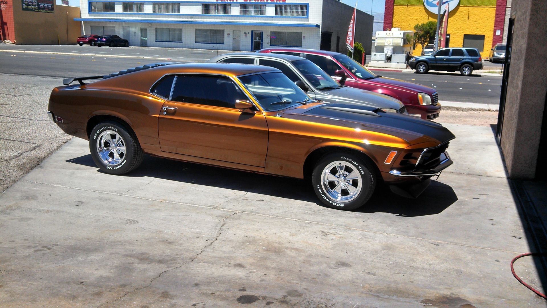 Orange classic muscle car parked in a lot, with other cars behind it and buildings in the background.