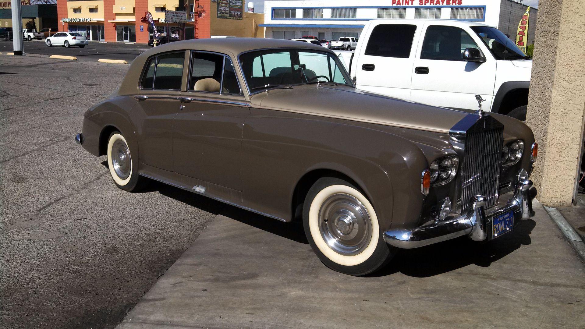 Brown vintage coupe parked on a street, with a white pickup truck in the background.