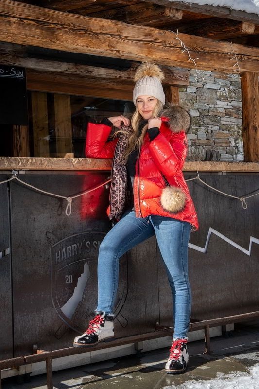 Frau in weißen Stiefeln, silberner Jacke und Jeans sitzt auf einem Stuhl neben dem Kamin im roten Zimmer.