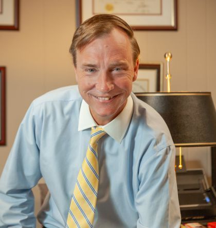 Smiling man in a light blue shirt and yellow striped tie in an office, leaning toward the camera.