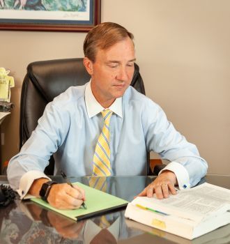 Man seated at a desk writing in a notebook beside an open book, wearing a light blue shirt and yellow striped tie.