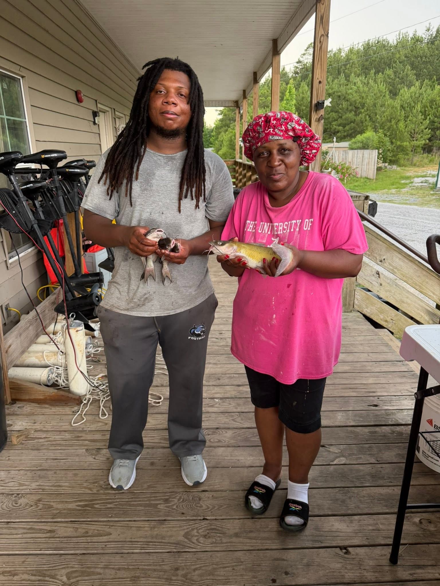Two people stand on a porch holding fish, with bicycles and a railing nearby.