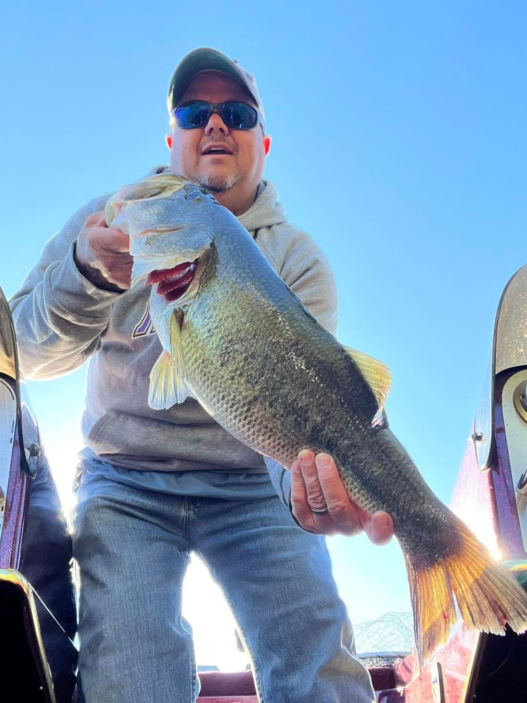 Angler holding a large fish on a boat, wearing a cap and sunglasses under a blue sky