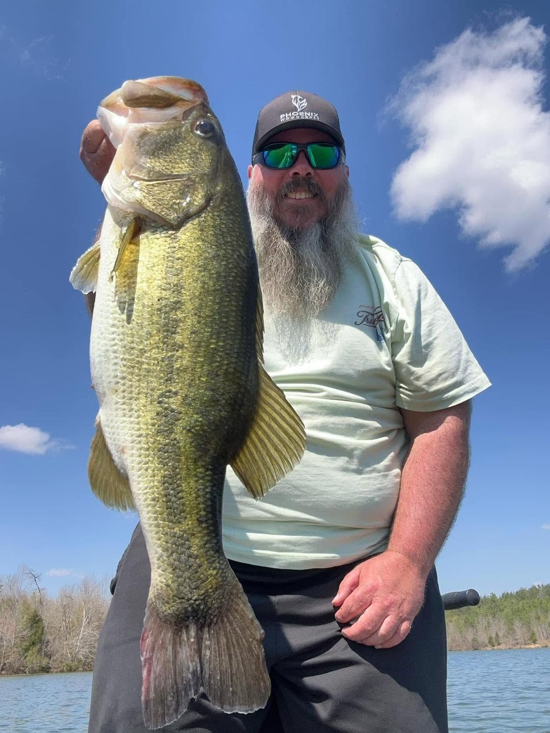 Man holding a large bass by a lake under a blue sky.